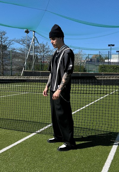 Camiseta deportiva negra con rayas blancas y pantalones negros, combinada con un gorro negro y zapatos negros con acentos blancos, en una cancha de tenis.