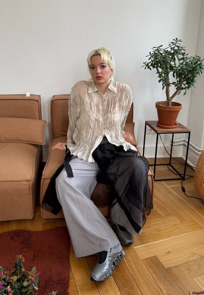 Camisa abotonada de color beige claro y arrugado; falda negra de capa transparente; pantalones de pierna ancha grises; zapatillas deportivas plateadas. Entorno interior con planta y sofá.