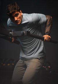 Vyras su tatuiruotu ranka ir išmaniuoju laikrodžiu atlieka dipus ant paralelinių barų prastai apšviestoje sporto salėje.