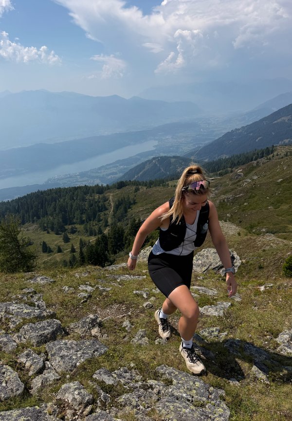 Femme faisant de la randonnée en montée sur un sentier montagneux rocheux avec une forêt et un lac en arrière-plan sous un ciel partiellement nuageux.