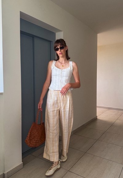 Mujer con top blanco sin mangas, pantalones anchos de cuadros beige, zapatos blancos con cordones, gafas de sol y llevando un bolso tejido marrón en el interior.