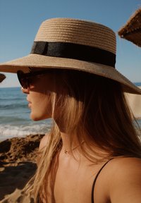 Grote strohoed met een zwarte riem, schuin gedragen; lang, blond haar; zonnebril; zandstrand en oceaan op de achtergrond.