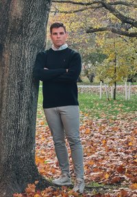 Άτομο που στηρίζεται σε ένα δέντρο, φορώντας μαύρο πουλόβερ, ανοιχτό γκρι παντελόνι και γκρι αθλητικά παπούτσια. Το φόντο περιέχει φθινοπωρινά φύλλα.