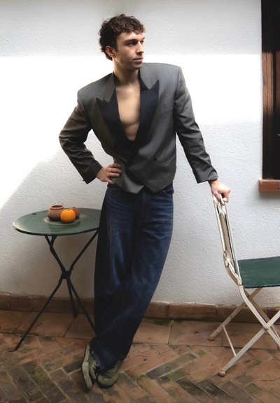 Joven con blazer oscuro abierto y vaqueros se apoya en una silla plegable blanca junto a una mesa redonda con macetas de barro y una naranja.