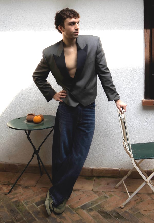 Jeune homme en veste sombre ouverte et jean, appuyé sur une chaise pliante blanche à côté d'une table ronde avec des pots en terre cuite et une orange.