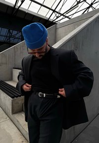 Μαύρο σακάκι και πουκάμισο συνδυασμένα με κομμένα μαύρα παντελόνια. Μπλε πλεκτό σκουφί, ζώνη με ασημένια αγκράφα, στέκεται σε μινιμαλιστικό σκηνικό από τσιμέντο.