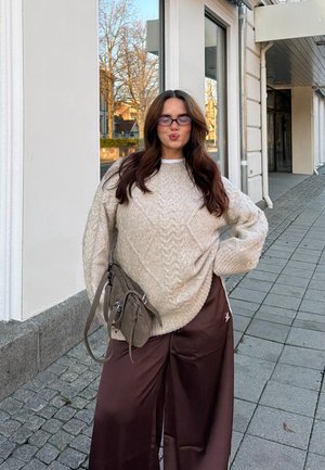 Suéter tejido de color beige claro con un patrón de rombos, combinado con pantalones anchos color marrón oscuro; un bolso gris apoyado contra el muslo.