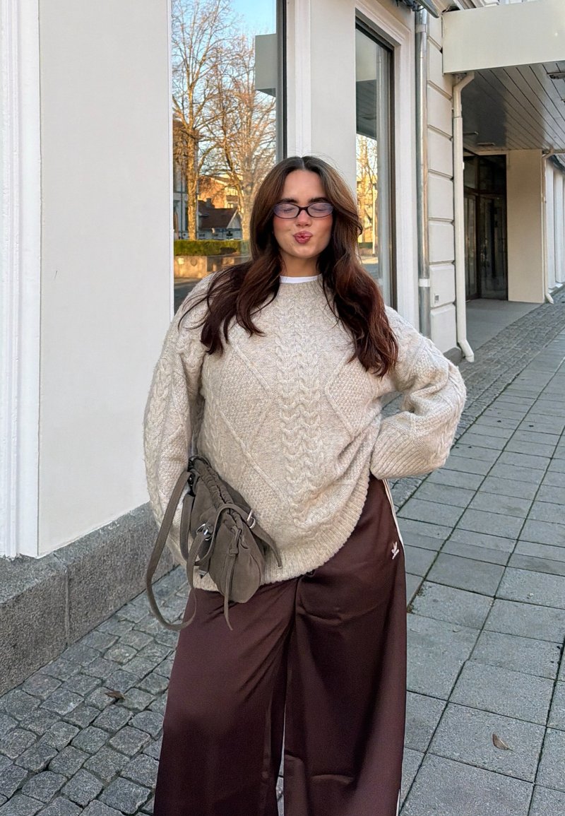 Strikket, lys beige genser med et diamantmønster, kombinert med mørkebrune vide bukser; en grå håndveske hviler mot låret.