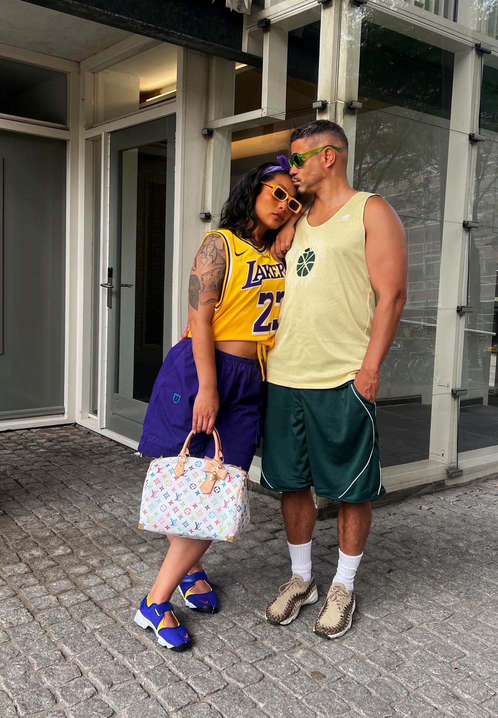 A woman is wearing a yellow basketball jersey, purple shorts, and sunglasses, while holding a multicoloured Louis Vuitton bag. A man is dressed in a light yellow tank top, green shorts, and patterned shoes.