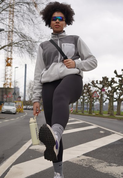 Chaqueta de forro polar gris y blanca con cremallera, leggings negros y calcetines estampados. Sosteniendo una botella de agua de color claro, usando gafas de sol reflectantes.