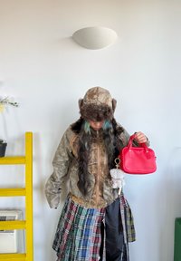 Kamuflasjjakke med fuskepelskant, rutete skjørt i grønn, blå og rød, holder en liten rød veske med en nøkkelring. Gul stige i bakgrunnen.