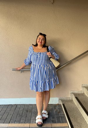 Vestido a rayas azul y blanco con mangas cortas abullonadas y dobladillo con volantes; combinado con sandalias en blanco y negro y un bolso de mimbre. Textura suave.
