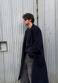 Manteau d'extérieur bleu marine à la coupe décontractée, porté par-dessus un pull foncé. Jean gris et lunettes de soleil rectangulaires complètent le look. Textures de tissu lisses.