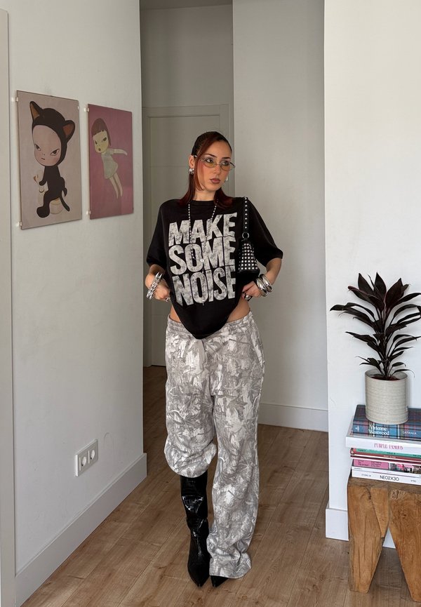 Νεαρή γυναίκα που φοράει μαύρο t-shirt με το σύνθημα "ΚΑΝΕ ΘΟΡΥΒΟ", παντελόνι με σχέδια, μαύρες μπότες και γυαλιά, στέκεται σε έναν μοντέρνο χώρο με έργα τέχνης και φυτά.