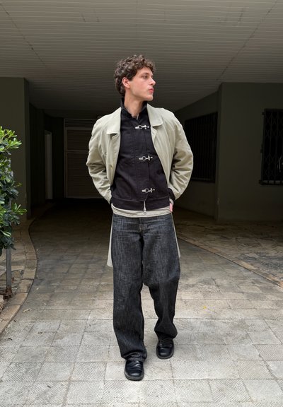 Joven caminando con las manos en los bolsillos, vestido con abrigo beige sobre chaqueta negra, vaqueros oscuros y sueltos, y zapatos negros en un pasillo al aire libre.