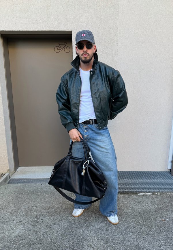 Un homme vêtu d’une veste en cuir noire, d’une casquette et de lunettes de soleil pose près d’une porte, en tenant un grand sac de voyage noir.