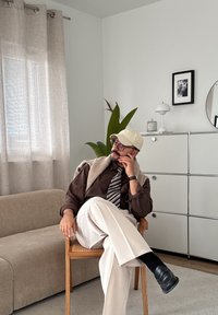 Mężczyzna w brązowej koszuli, w paskowaną krawat, beżowych spodniach i czapce, siedzący w pozycji skrzyżowanej na drewnianym krześle w nowoczesnym salonie z rośliną i szafką.