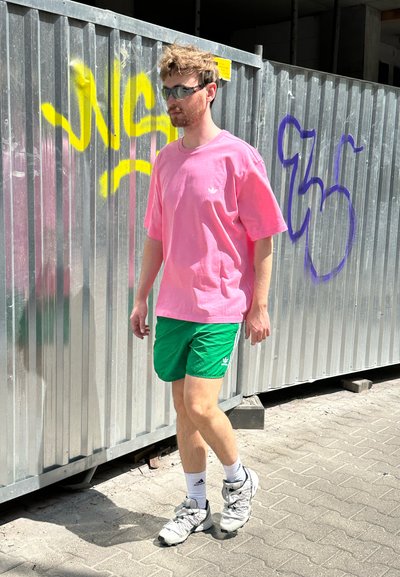 Camiseta de algodón rosa, pantalones cortos verdes, zapatillas grises y calcetines blancos. El fondo presenta una pared de metal corrugado con graffiti.