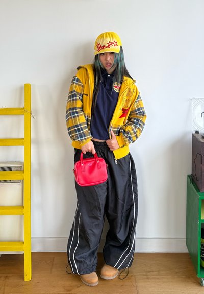 Chaqueta de cuadros amarilla sobre una camisa azul marino, pantalones deportivos negros con rayas blancas, bolso rojo y gorra amarilla con cinco estrellas. Calzado marrón.