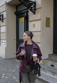 Casaco de pele sintética burgundy com metais prateados sobre um suéter castanho, combinado com calças de padrão de leopardo e uma bolsa preta. Segurando uma chávena de café.