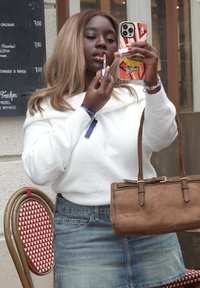 Hvit off-shoulder genser, denimskjørt og brun semsket skinnveske. Hun holder et leppestiftetui som reflekteres i et mobildeksel med sportstema.