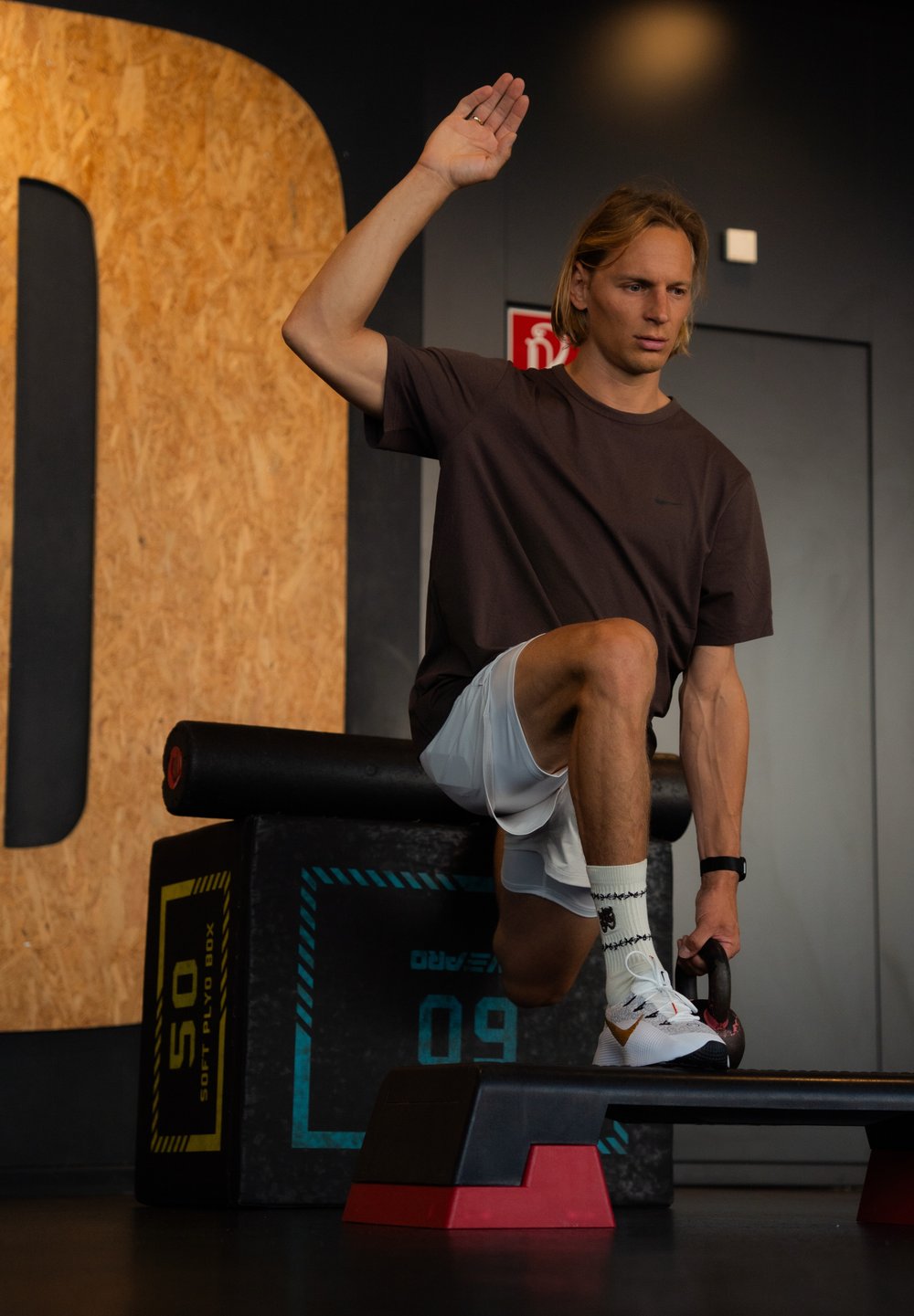 Melnā un sarkanā kettlebell, kuru tur cilvēks uz melnas plyo kastes, tērpts brūnā krekla un baltās šortos, sporta zālē ar teksturētu sienu.