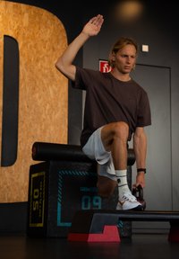 Melnā un sarkanā kettlebell, kuru tur cilvēks uz melnas plyo kastes, tērpts brūnā krekla un baltās šortos, sporta zālē ar teksturētu sienu.