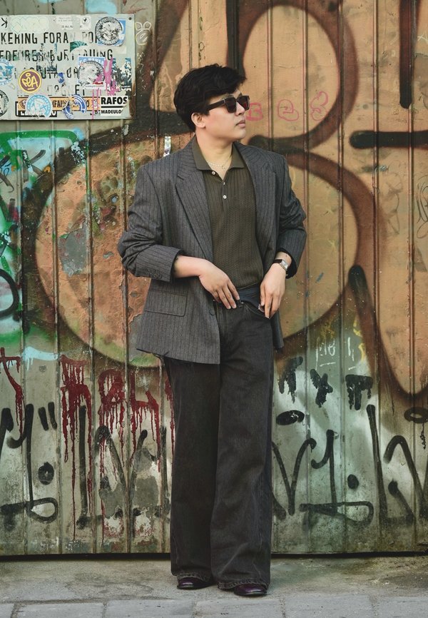 Personne portant des lunettes de soleil, un blazer gris à fines rayures et un pantalon foncé, debout contre un mur couvert de graffiti avec des autocollants.