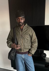 Veste bomber vert olive avec fermeture éclair frontale, poignets côtelés et une poche zippée, accompagnée d'un jean bleu ample et d'un sac à bandoulière noir.