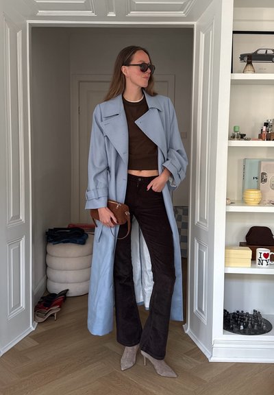 Mujer con abrigo largo azul claro, top corto marrón oscuro, pantalones negros, gafas de sol, sosteniendo un bolso marrón, de pie en la puerta de una habitación con estanterías.