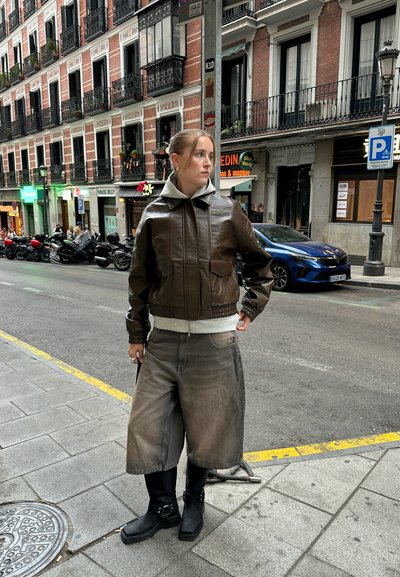 Chaqueta de cuero marrón corta sobre una sudadera gris, combinada con pantalones cortos de mezclilla grises anchos y botas negras gruesas. Entorno urbano.