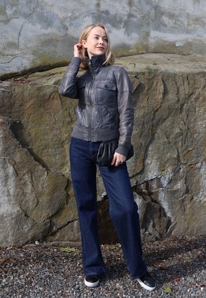 Mujer con chaqueta de cuero gris y vaqueros oscuros está sobre grava frente a una gran pared de roca texturizada, sosteniendo un bolso de mano negro.