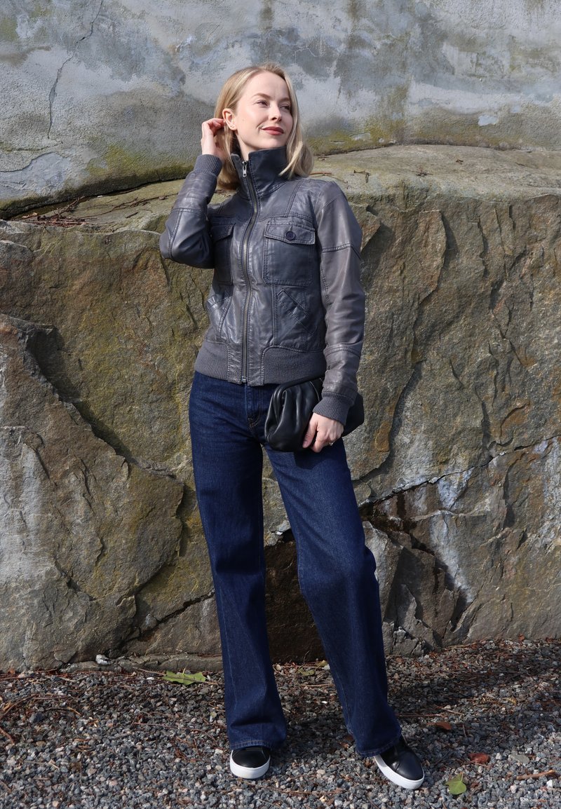 Femme en veste en cuir grise et jean foncé se tient sur du gravier devant un grand mur de roche texturée, tenant une pochette noire.