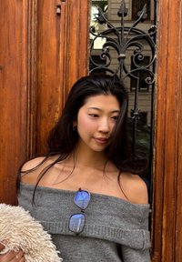 Junge Frau in grauem schulterfreiem Pullover, die einen beigen, flauschigen Gegenstand hält, steht vor einer Holztür mit dekorativer schwarzer Schmiedearbeit.