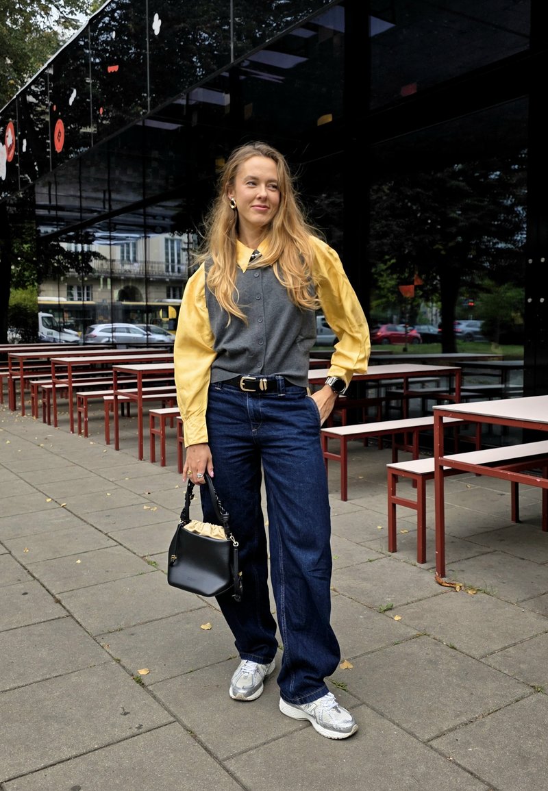 Gilet gris sur une chemise jaune à manches longues, associé à un jean bleu ample et des baskets argentées. Elle tient un petit sac à main noir.