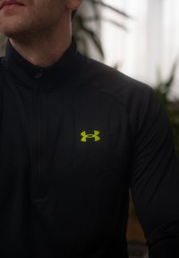 Haut de sport noir zippé avec un col haut, arborant un logo Under Armour vert sur la poitrine gauche. Texture lisse, design ajusté.