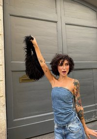 Ung tatuerad kvinna i jeanskläder som lyfter en svart lurvig väska, står framför en stor grå träport.