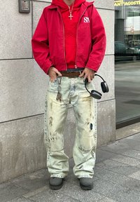 Κόκκινο φούτερ με φερμουάρ πάνω από μια κόκκινη μπλούζα φούτερ, συνδυασμένο με ανοιχτό μπλε φθαρμένα τζιν με τρύπες. Μαύρα ακουστικά κρέμονται από τον βρόχο της ζώνης.