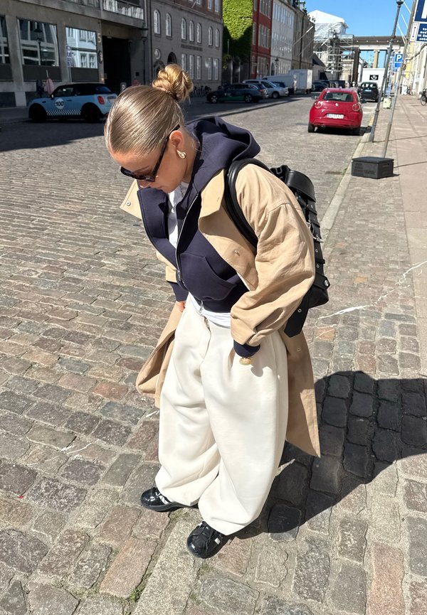 Femeie cu ochelari de soare, pardesiu bej, hanorac bleumarin, pantaloni albi largi și pantofi negri, stând pe o stradă pietruită însorită cu mașini parcate în apropiere.