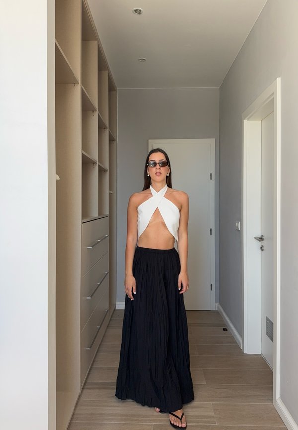 Femme portant un crop top blanc à col halter, une longue jupe noire, des lunettes de soleil et des sandales, debout dans un couloir minimaliste avec un placard et des portes blanches.