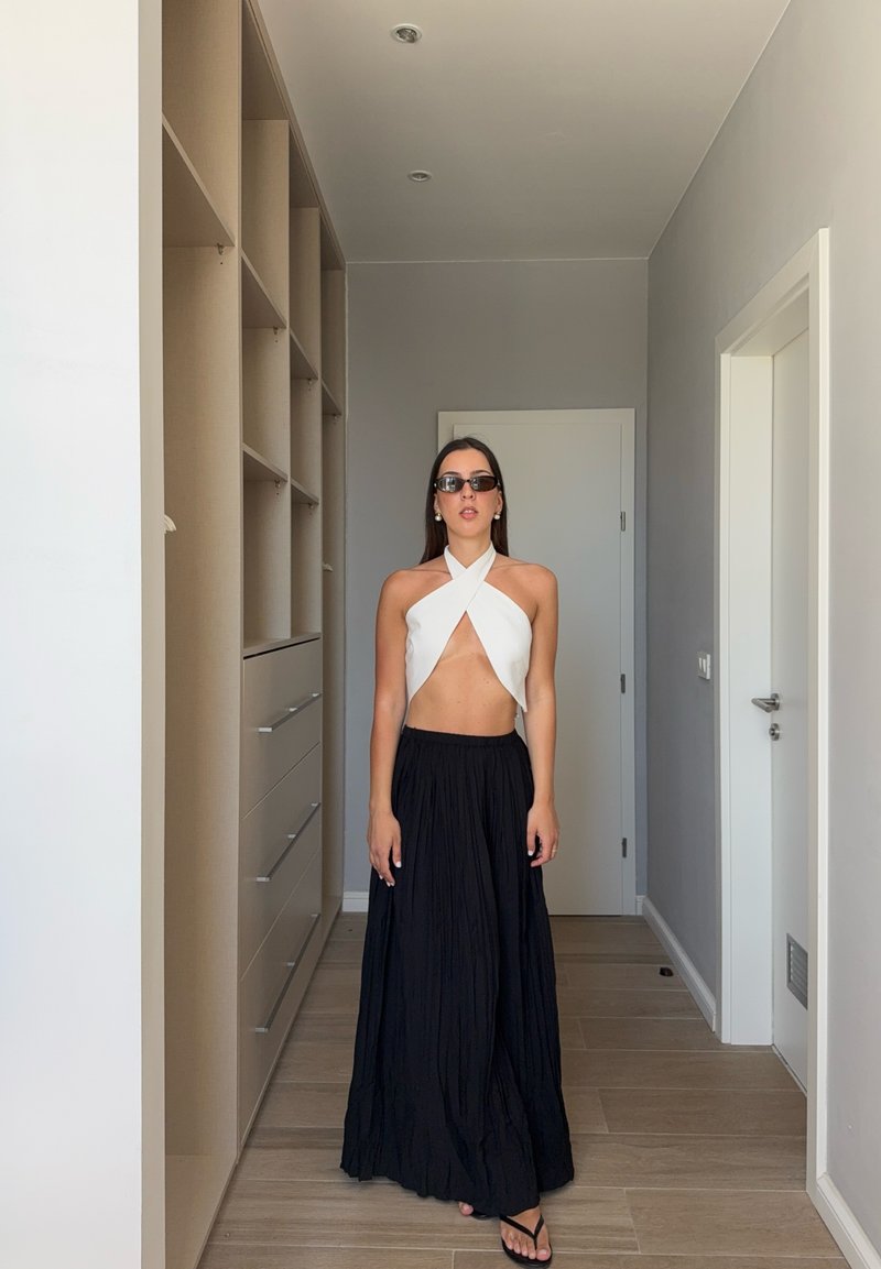 Femme portant un crop top blanc à col halter, une longue jupe noire, des lunettes de soleil et des sandales, debout dans un couloir minimaliste avec un placard et des portes blanches.