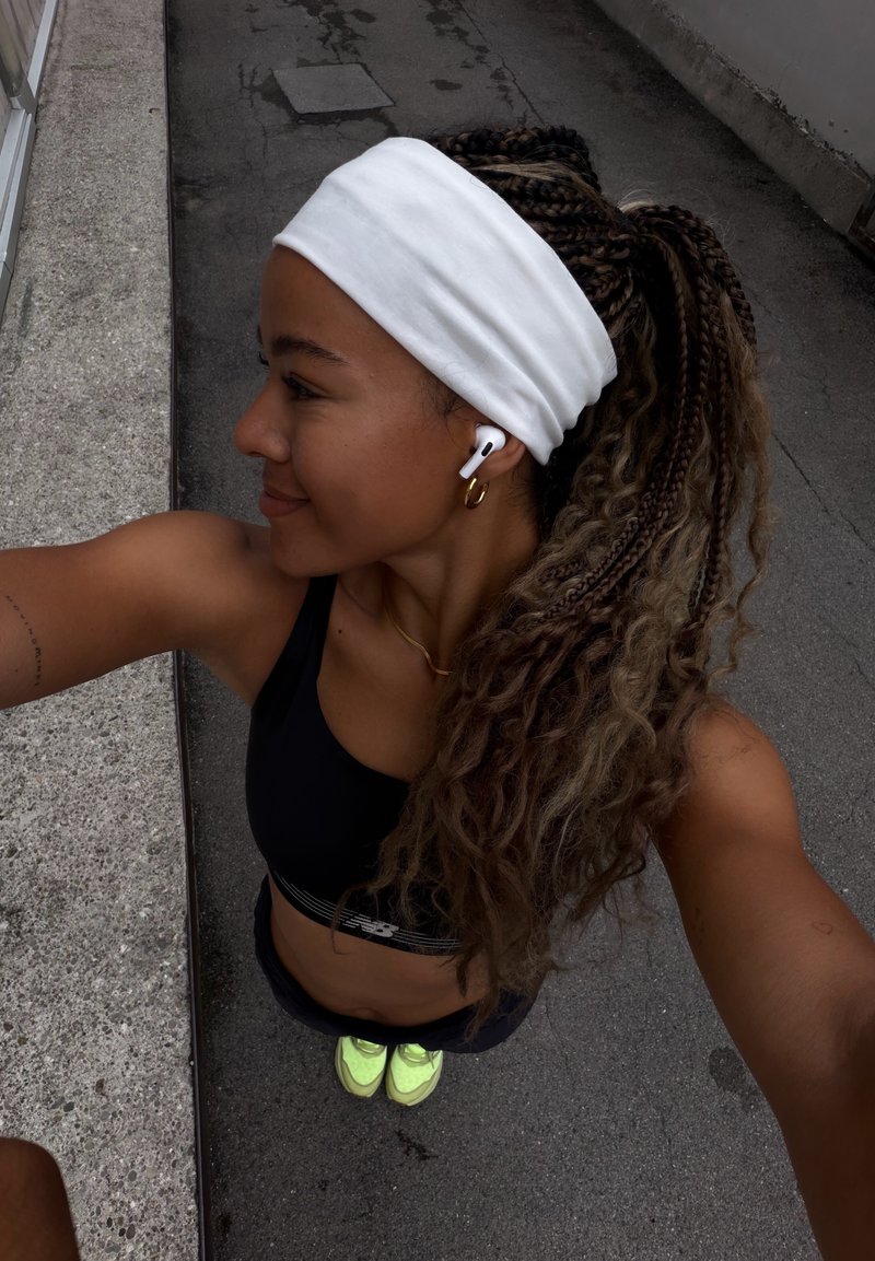 Jeune femme aux cheveux tressés et bandeau blanc, portant des vêtements de sport et des écouteurs, debout à l'extérieur sur un trottoir à côté d'un rebord en béton.
