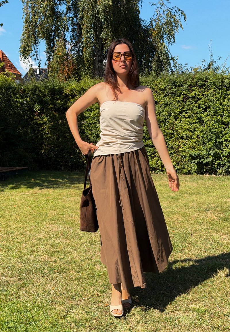 Woman wearing strapless white top, brown skirt, and orange sunglasses walking on grass with bag in hand under clear sky.