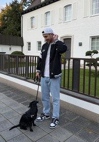 Schwarze Collegejacke mit weißen Streifen, weißes T-Shirt, hellblaue Jeans und schwarze Nike-Turnschuhe. Ein schwarzer Hund an der Leine sitzt neben ihm.