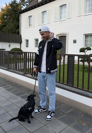 Chaqueta de béisbol negra con rayas blancas, camiseta blanca, jeans azul claro y zapatillas Nike negras. Un perro negro con correa está sentado a su lado.