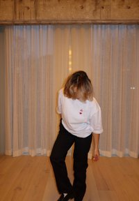 T-shirt bianco a maniche corte con un cuore rosso e il logo "A", abbinato a pantaloni neri. Pavimento in legno chiaro e tende trasparenti sullo sfondo.