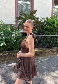 Jauna moteris ruda be rankovių suknele, laikanti rankinę, stovi akmeniniu taku su žaliais krūmais ir pastatu fone.