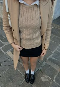 Béžový pletený svetr s kabelkovým vzorem, černá mini sukně, bílé kotníkové ponožky a černé loafers, doplněné béžovým kabátem.
