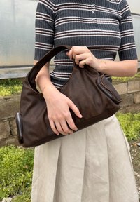 Personne tenant un grand sac à main en cuir marron avec des fermetures éclair latérales, portant un haut rayé gris et une jupe beige, debout à l'extérieur près d'un mur en pierre et de la verdure.