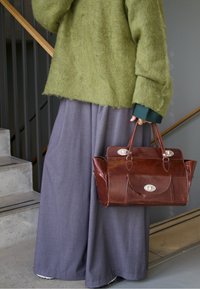 Hnědá kožená kabelka se stříbrnými kovovými doplňky, držená osobou v zeleném chlupatém svetru a šedých širokých kalhotách.